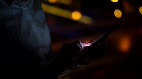Young woman stay against window, texting using mobile phone, closeup shot in dark room. Blurred background, dark city traffic lights on road seen outdoors