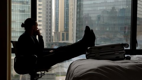 Thoughtful man rest at dark hotel room, stare to city, sitting in chair against window. Traveller stretch out legs on suitcase lying on bed, put chin on hand. Silhouetted slow motion shot