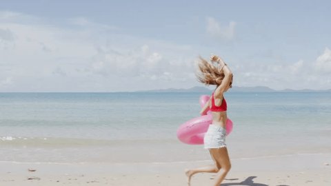 Tracking shot of young woman running on the beach holding a pink inflatable flamingo. Slow motion of woman having fun on the beach. 