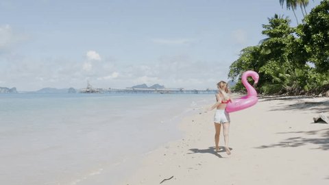 Young woman making follow me gesture on the beach. Tracking shot of a woman holding an inflatable flamingo and running on the beach. Slow motion 