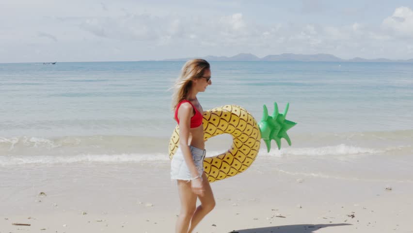Woman walking on empty beach holding inflatable pineapple enjoying summer vacation on tropical beach holiday wearing a bikini. Tracking shot 