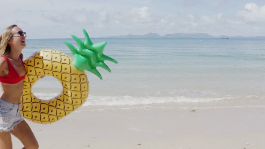 Woman walking on empty beach holding inflatable pineapple enjoying summer vacation on tropical beach holiday wearing a bikini. Tracking shot 