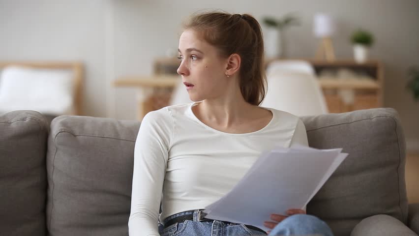 Sad woman sitting on couch at home reads received bad news holds documents paper letter feels desperate about financial problems, domestic bills or debt, girl student worried college expulsion concept | Shutterstock HD Video #1028517131