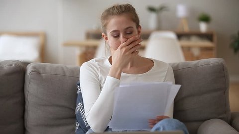 Sad woman sitting on couch at home reads received bad news holds documents paper letter feels desperate about financial problems, domestic bills or debt, girl student worried college expulsion concept