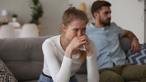 Millennial couple in quarrel sitting apart on couch focus on crying unhappy girl holding handkerchief feels desperate, break up divorce, cheating and problems in relationship crisis, abortion concept