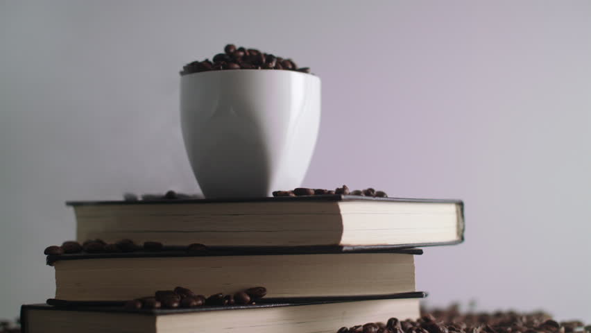 White coffee mug, surrounded by roasted coffee beans on a stack of books, rotating on a display turntable. With steam smoke and falling beans.