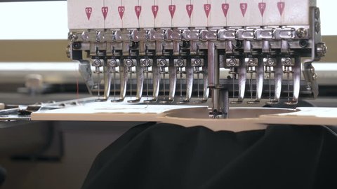 In slow motion video in a garment factory, a sewing machine embroiders various companies and engravings. Concept of: Automated work, Sewing machine, Engraving, Thread, Fabric, Needle.