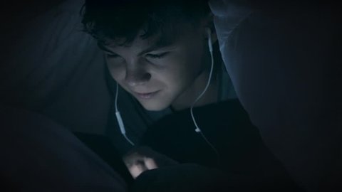 Night shot of teen boy with headphones using tablet pc under blanket. Happy child playing with cellphone or smartphone on a bed. Light reflection on face cute young boy. 