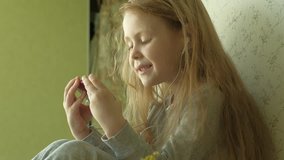 beautiful little girl with red hair in gray pajamas sitting on the window with headphones is using a phone, watching a video or playing and laughing, close-up portrait