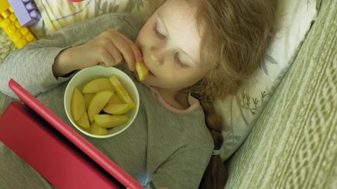beautiful little girl with red hair lying on the sofa uses a computer tablet and eats apples in the interior, around the toys are scattered pink, purple and yellow