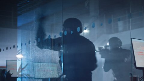 Masked Squad of Armed SWAT Police Officers Storm a Dark Seized Office Building with Desks and Computers. Soldier Breaks a Glass with His Arm and Team Continue to Move. Warm Color Grade.