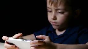 A child watching a cartoon at home at night, using a smartphone, sits at a table lit by a lamp, close-up with a focus first on his hands and then on his face