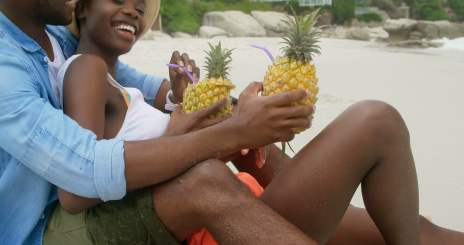 Side view of African american couple interacting with each other on the beach. They are drinking pineapple juice.