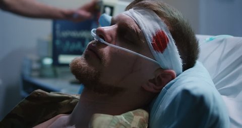 Close up shot of man lying in hospital bed with bloody bandage around his head while hand setting up device on the background