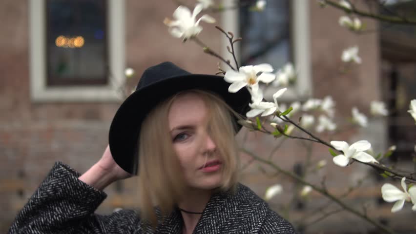 young woman with long hair in a gray long coat with a hat at sunset among flowering trees