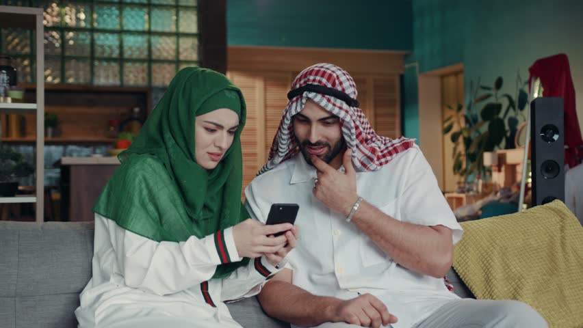 Arab cheerful couple at home sitting in traditional dress using mobile phone. Surfing the internet, social networks, being online. Information, gadgets and devices. Technology concept