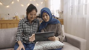 at cozy bright home muslim woman make video phone call to family introduce asian friend while two girls relaxing at home kitchen sit on couch. happy multi friendship waving hands talking to tablet