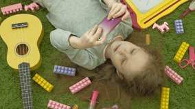 beautiful little girl with red hair lying on the floor on a green background in headphones and using a phone, watching a video or playing, top view, next to them are toys of yellow, pink and purple