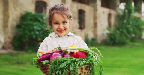 Slow motion of happy little girl is holding a basket with fresh biologic just harvested vegetables and smiling in camera on a background of a countryside farm.Vegetarian people