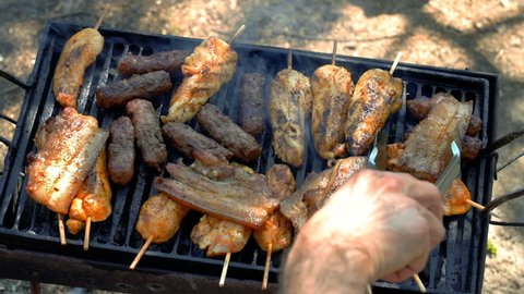 Hand Using Kitchen Tongs for Grilling Meat on the Barbecue Grill - Slow Motion. Assorted Delicious Grilled Meat Over the Coals on a Barbecue. Outdoor Roasting on a Grill. Barbecue Party.