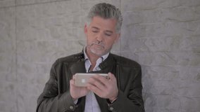 Serious mature man using mobile phone. Handsome focused grey haired man standing near wall, shaking head and thinking while using smartphone. Technology concept