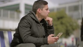 Thoughtful man using cell phone outdoor. Concentrated young man sitting on bench, shaking head and looking away while texting by smartphone. Technology concept