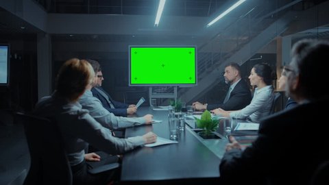 Late at Night In Corporate Meeting Room: Board of Directors, Executives and Businesspeople Sitting at Negotiations Table, Talking and Using Green Mock-up Screen Wall TV for Video Conference Call.