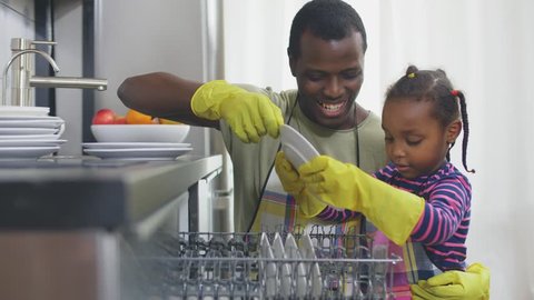 Afro-American father and little daughter kneeling next to dishwasher in bright kitchen