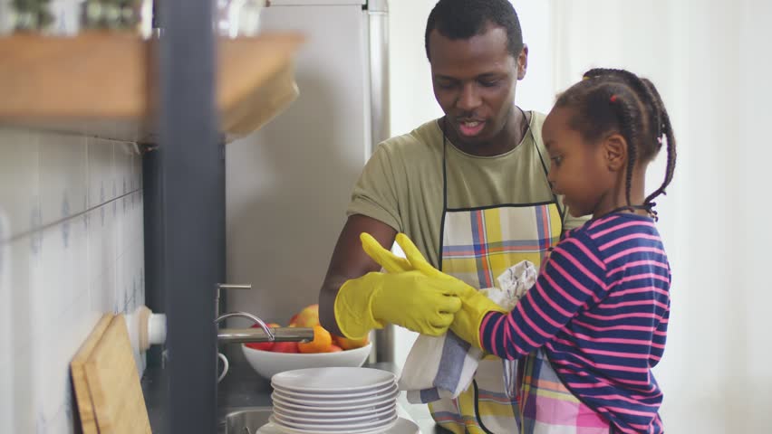 Black man and his little daughter in yellow rubber gloves wiping plates in modern kitchen Royalty-Free Stock Footage #1028621489