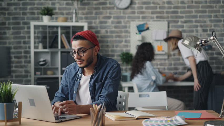 Cheerful young man hipster wearing hat and glasses is using laptop typing working in modern loft style office enjoying modern technology. Youth and work concept. Royalty-Free Stock Footage #1028622962