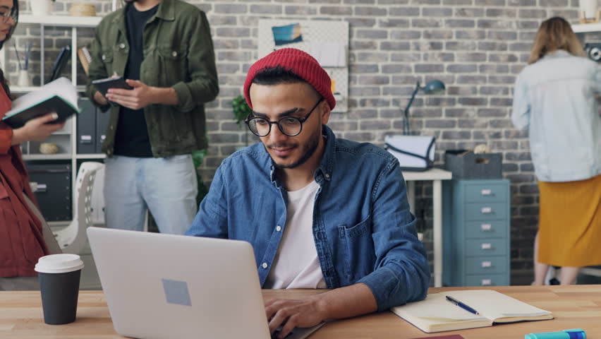 Zoom out time-lapse of male employee handsome hipster in trendy clothing working with laptop busy with project in office. Coworkers are moving around. Royalty-Free Stock Footage #1028623136