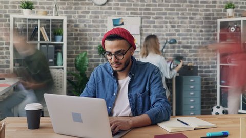Zoom out time-lapse of male employee handsome hipster in trendy clothing working with laptop busy with project in office. Coworkers are moving around.