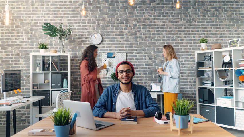 Zoom in time-lapse of happy business owner looking at camera and smiling sitting in office while men and women are moving around working. Millennials and job concept.