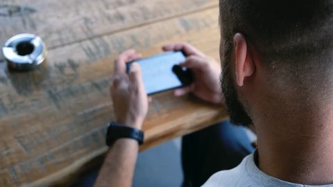 A young man with a beard playing mobile games on the phone in an open-air cafe. Close up