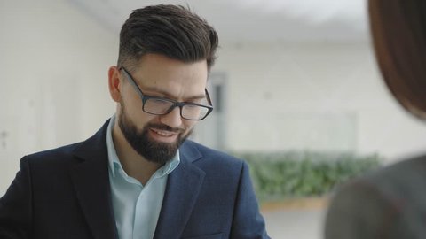 Young friendly business man in glasses and a suit communicates with a colleague and smiles. Business solid man with a beard says something to a woman. Shot over the shoulder of a woman