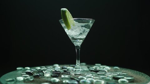 Rotating martini glass with a slice of lemon and ice blocks, rotating on a beautiful plateau with shiny stones, shot on a black infiniy background.