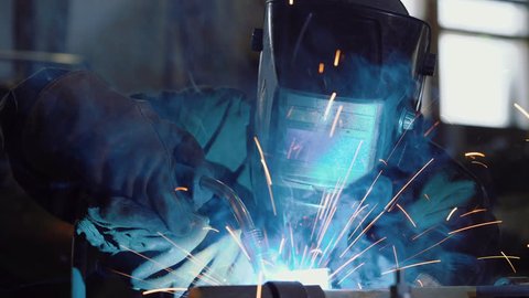 Close up: worker in face shield or protective mask uses gas welding torch to connect the seam of metal part in industrial factory. Blue fire flickers, orange sparks are flying, blacksmith manual work.