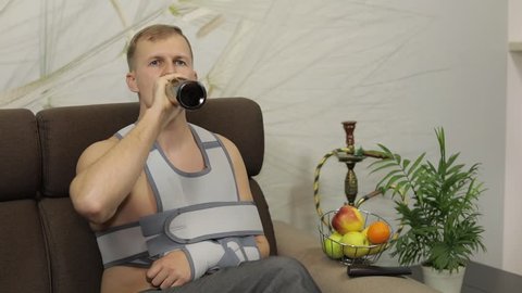 Man with shoulder injury. Painful, bored man with a broken arm wearing arm brace sitting on a sofa at home and drinks beer. Patient in a bandage for fixing of an elbow joint and a humeral belt