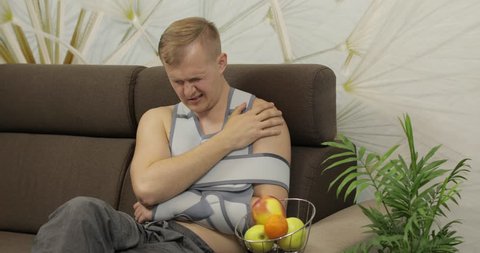 Man with shoulder injury. Painful, bored man with a broken arm wearing arm brace sitting on a sofa and eating apple at home. Patient in a bandage for fixing of an elbow joint and a humeral belt