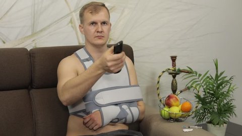 Man with shoulder injury. Painful, bored man with a broken arm wearing arm brace sitting on a sofa at home watching TV. Patient in a bandage for fixing of an elbow joint and a humeral belt
