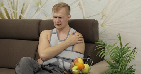 Man with shoulder injury. Painful, bored man with a broken arm wearing arm brace sitting on a sofa and watching TV at home. Patient in a bandage for fixing of an elbow joint and a humeral belt