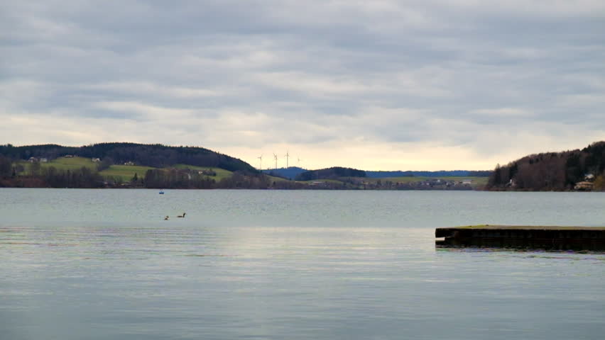 Wide angle shot of Lake Mattsee in Austria with birds swimming across the water in foreground and fast spinning wind turbines in the background