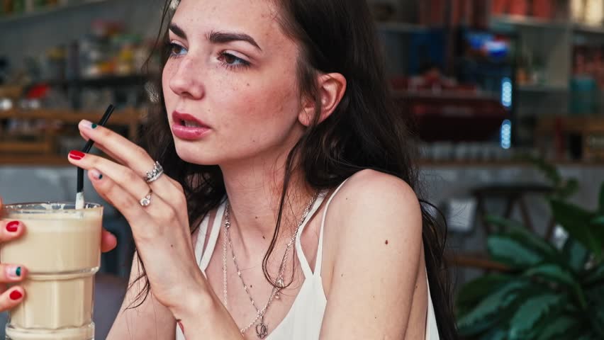 Contemporary beverage consumption: stylish young female with long dark hair sips creamy beige liquid through black straw in trendy indoor establishment, conveying leisure and contentment with relaxed shoulders and confident posture. She wears sleeveless cream-colored tank top layered with delicate silver chain necklaces, statement rings decorating manicured hands featuring vibrant red and navy blue nail polish. Fluid motion unfolds elegantly, beginning with subject holding tall glass at mid-frame while appearing contemplative, transitioning through sipping action with straw positioned at lips, concluding with serene moment of enjoyment as beverage rests closer to body. Soft warm illumination bathes scene with golden and amber tones, creating intimate atmosphere that highlights facial features and shoulder details against sophisticated cafe or cocktail bar background. Blurred warm-toned hardwood furniture, shelving with ambient blue neon lighting visible on right side, various bottles, decorative elements softly out of focus, and lush green potted plants provide natural accents through bokeh effect that enhances depth. Composition employs shallow depth of field with subject positioned slightly off-center in three-quarter profile view, capturing both expression and ritual of liquid consumption within color palette featuring cream, beige, warm golds, contrasting red and blue nail polish, silver metallic jewelry, white garment, and muted interior tones. This footage demonstrates natural candid movement with smooth temporal pacing that visualizes relaxation, sophistication, and casual social engagement suitable for millennial and Gen-Z demographics.