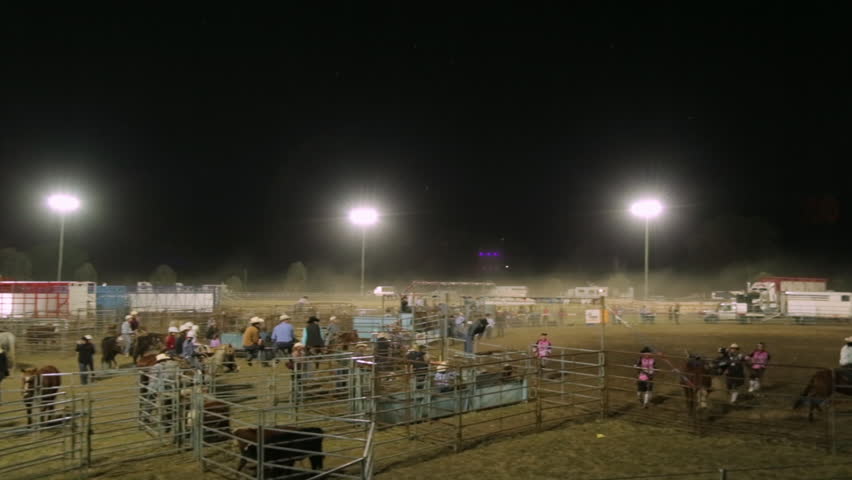 A panning long shot of a bull going on a solo rampage as people tries to tame it and some people in the stands watch on.