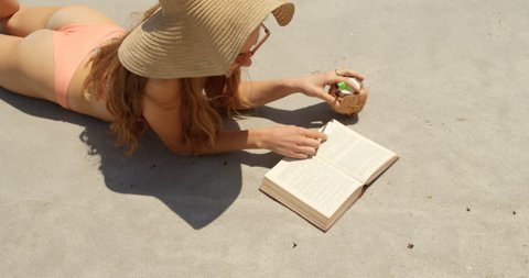 High angle view of Caucasian woman in hat reading a book on the beach. She is drinking coconut water