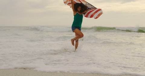Front view of African American woman with waving American flag dancing on the beach. She is having fun
