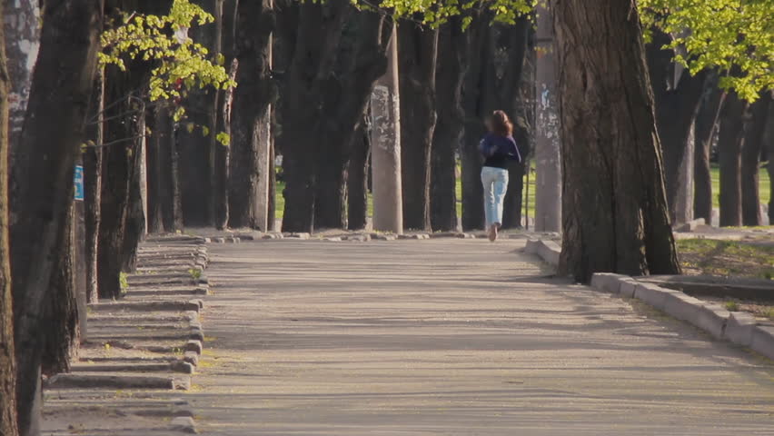 A young girl in jeans and a blouse runs along the avenue next to the roadway with green trees in the spring in the city