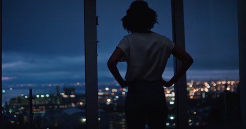 young woman looking out window enjoying view of city at night contemplating successful lifestyle planning ahead on calm urban evening