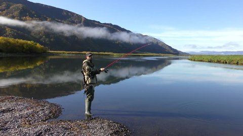 Fishing by means of spinning on the rivers on the Kamchatka Peninsula. Russia, September 2018. A man with a carbine behind his back caught a fish on a spinning in the river.