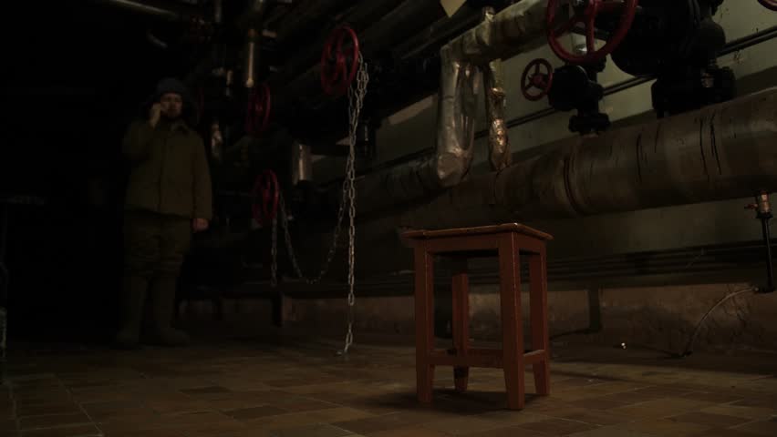 A man with a smartphone and in boots is walking in front of the camera. Tiled floor, many pipes, basement with dim light. A light in the end of a tunnel.